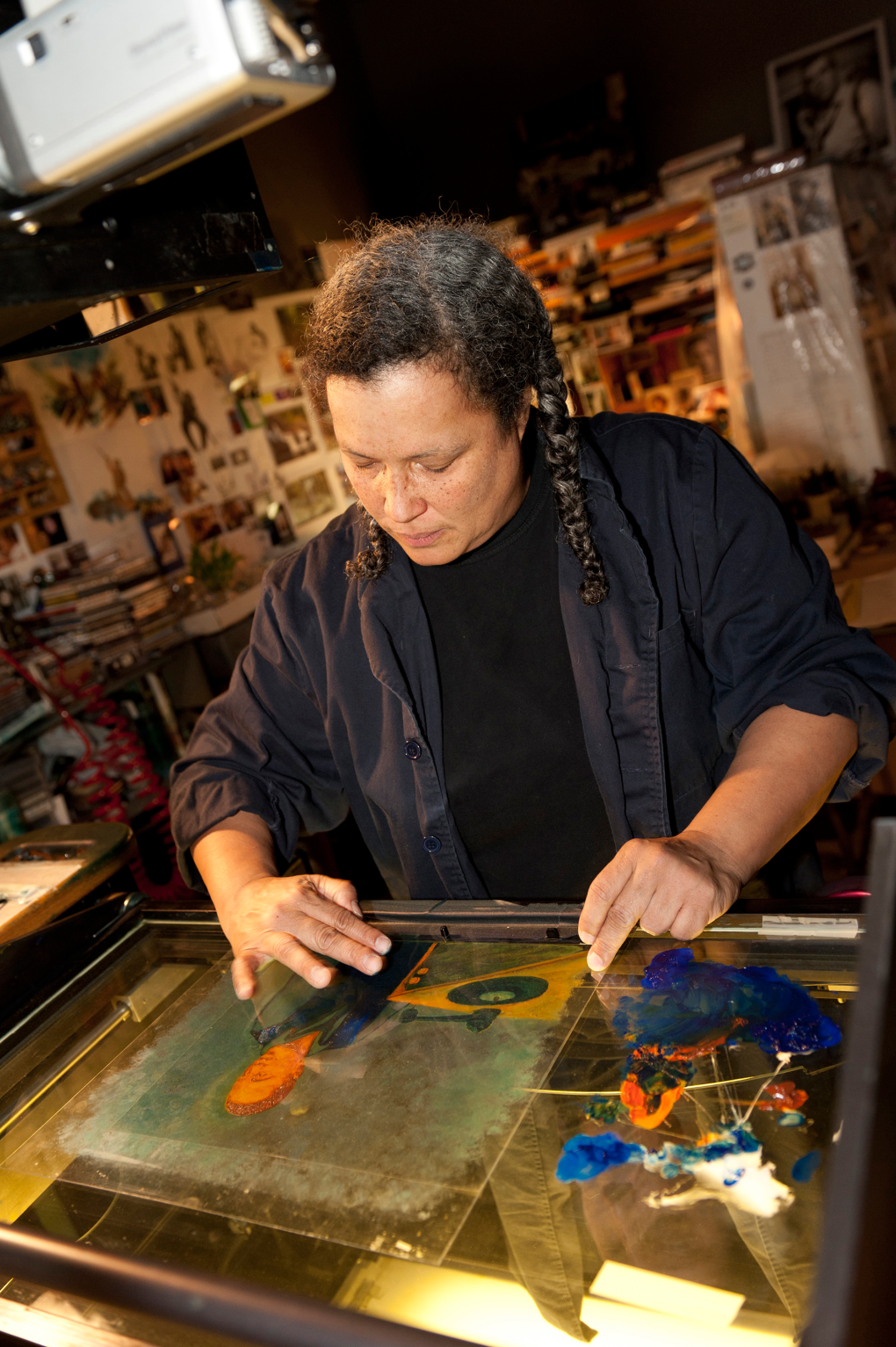 A woman with long braided hair works intently on a glass multiplane, using their hands to manipulate colorful paint in an art studio filled with tools and supplies.