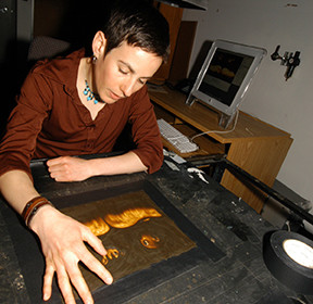 A woman with short brown hair wearing a brown shirt works clay on a backlit surface